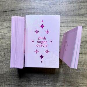 May include: A pink deck of oracle cards with the text "pink sugar oracle" on the front. The cards are stacked in a pile and the top card is visible.