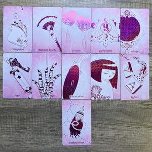 May include: A set of 11 pink and white cards with glitter accents. Each card features a different image and text. The images include a coin purse, makeup brush, pasties, planchette, mirror, key, chicken feet, pizza cutter, kokeshi doll, lighter, and rabbit's foot.