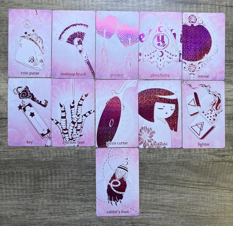 May include: A set of 11 pink and white cards with glitter accents. Each card features a different image and text. The images include a coin purse, makeup brush, pasties, planchette, mirror, key, chicken feet, pizza cutter, kokeshi doll, lighter, and rabbit's foot.
