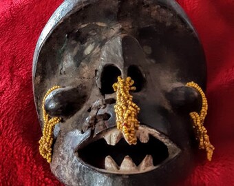 May include: A wooden mask with a carved face, open mouth, and large teeth. The mask is painted black and has yellow beads around the eyes and nose.