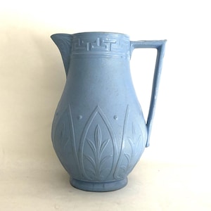Peut inclure: Une cruche en céramique bleu clair présentant une surface texturée avec un motif de feuilles répétitif autour de la base et un motif géométrique grec entourant le bord supérieur. Le récipient comprend un bec verseur et une anse angulaire.
