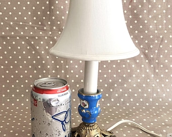 May include: A small, decorative table lamp with a white lampshade and a blue and gold base. A can of Club Soda is next to the lamp. The lamp has a white cord and a white bulb. The can is 12 fl oz.