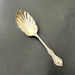 May include: A silver serving spoon with a shell-shaped bowl and a decorative handle. The spoon is made of a shiny, reflective metal and is designed for serving food. The spoon is likely vintage or antique.