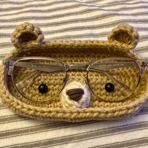Op de afbeelding: Een gehaakt brilletje in de vorm van een beer met een bruine neus en zwarte ogen. Het etui bevat een bril met heldere glazen en een zilveren montuur.