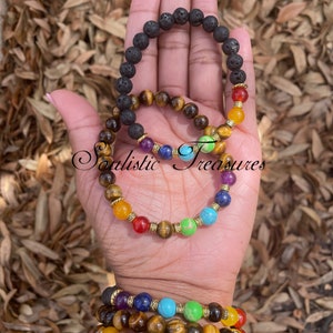 May include: A set of seven chakra bracelets featuring lava rock, tiger's eye, amethyst, lapis lazuli, turquoise, green aventurine, and red jasper beads. The bracelets are stacked on a person's wrist.