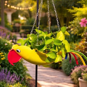 May include: A yellow parrot-shaped hanging planter filled with green plants. The planter has a red beak and colorful tail feathers. It hangs from a black metal pole with a chain, set against a garden backdrop with flowers and a stone path.