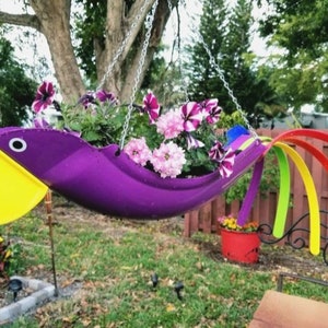 May include: A purple and yellow bird-shaped hanging planter with a rainbow tail. The planter is filled with pink and white flowers.