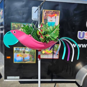 May include: A pink parrot-shaped planter with a turquoise beak and colorful tail feathers. The planter is suspended by chains from a metal rod and filled with green and red plants. The background is a black trailer.