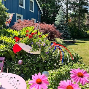 May include: A white metal bird planter with red, white, and blue accents. The planter is filled with colorful flowers and is hanging from a metal stand. The planter is in front of a blue house with a green lawn.