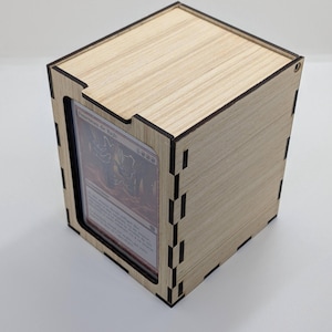 May include: A wooden Basic TopLoader Box with a card visible inside. The box is made of light-colored wood and has a rectangular shape. The text "Basic TopLoader Box" is at the top. The box is designed to hold trading cards. The text "Kamsing" is at the bottom.