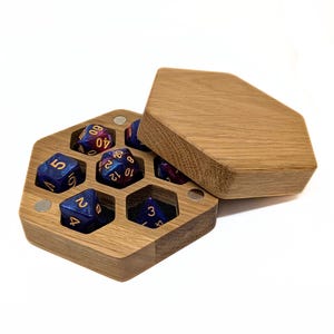 Peut inclure: Une boîte à dés hexagonale en bois avec un couvercle amovible, comprenant six compartiments individuels. À l'intérieur se trouvent plusieurs dés polyédriques aux tons bleu et violet avec des chiffres dorés. La boîte présente un grain de bois visible.