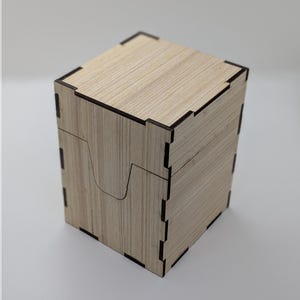 Peut inclure: Une boîte de rangement en bois avec le texte "BASIC DECKBOX" en orange. La boîte est en bois clair avec des bords marron foncé. La boîte est carrée avec un couvercle et une découpe incurvée sur le devant. Le texte "Kamsing" est en dessous.