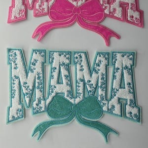 May include: Three embroidered patches with the word "MAMA" in white letters with a floral pattern on a pink, blue, and pink background. Each patch has a bow in a matching color.