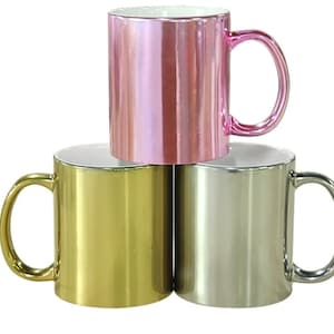 May include: Three ceramic mugs with metallic finishes. The mugs are stacked on top of each other. The top mug is pink, the middle mug is gold, and the bottom mug is silver.