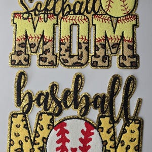 May include: Two gold glitter patches with black lettering and leopard print accents. The top patch says "Softball Mom" with a yellow softball. The bottom patch says "Baseball Mom" with a red baseball.