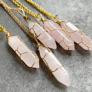Könnte beinhalten: Vier Halsketten mit blassrosa, halbtransparenten, doppelt endenden Kristallspitzen. Jeder Kristall ist in einen goldfarbenen Drahtkäfig gefasst und an einer passenden goldfarbenen Metallkette befestigt. Die Kristalle haben eine glatte, polierte Oberfläche.