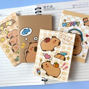 Könnte beinhalten: Vier Notizbücher mit Cartoon-Capybara-Illustrationen. Die Einbände zeigen beige Karomuster, einfarbig braune Hintergründe und bunte Grafiken mit Äpfeln, Vögeln, Herzen sowie den Schriftzügen "Capybara" und "Happy" in verspielten Schriftarten.