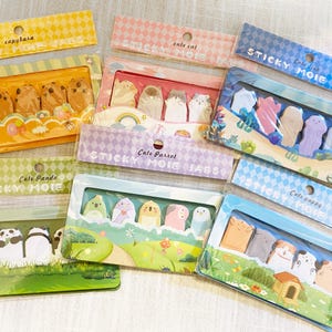 Könnte beinhalten: Eine Sammlung farbenfroher Haftnotiz-Sets mit verschiedenen niedlichen Tierdesigns, darunter Capybaras, Katzen, Papageien, Fische, Pandas und Welpen. Jedes Set ist in einer transparenten Kunststoffbox mit dekorativen Hintergründen und Textetiketten verpackt.