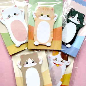 Könnte beinhalten: Eine Sammlung von katzenförmigen Haftnotizen in verschiedenen Farben, darunter Weiß, Orange, Braun und Schwarz. Jede Notiz zeigt ein niedliches Katzendesign mit ausgestreckten Pfoten und einem leeren Bereich zum Schreiben. Die Notizen sind in Klarsichtfolie verpackt.