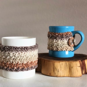 May include: Two mugs with crocheted cozy covers. The mugs are both white with a brown, beige, and light brown crocheted cover. The mug on the left is white and the mug on the right is blue.
