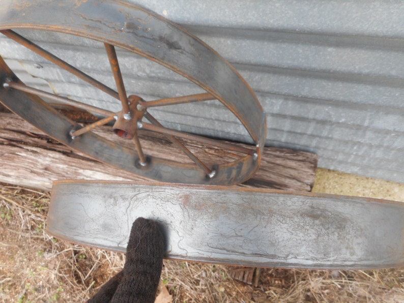 May include: Two metal wheel rims, one with spokes and one without. The rims are rusty and have a grey patina. The rim with spokes is lying on a piece of wood.