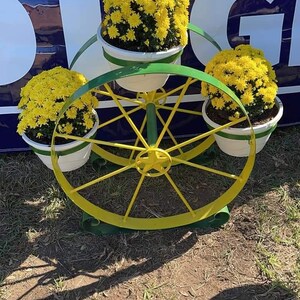Puede incluir: Un soporte de plantas de metal amarillo con un dise&ntilde;o de rueda. El soporte tiene tres macetas blancas con crisantemos amarillos.