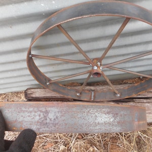 Puede incluir: Una rueda de metal oxidada con radios y una llanta de metal. La rueda est&aacute; apoyada sobre una superficie de madera. Hay una llanta de metal oxidada separada que est&aacute; apoyada en el suelo.