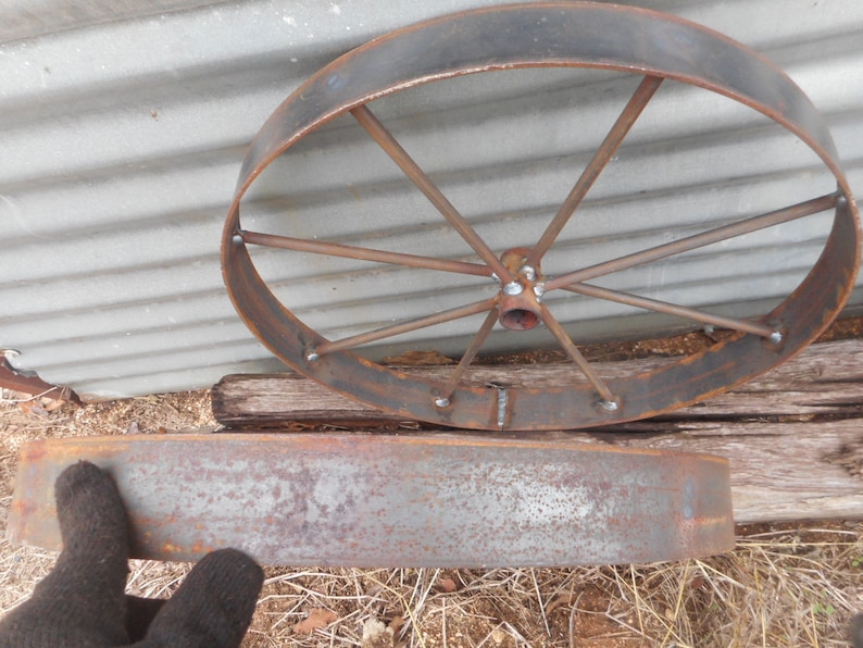 Puede incluir: Una rueda de metal oxidada con radios y una llanta de metal. La rueda est&aacute; apoyada sobre una superficie de madera. Hay una llanta de metal oxidada separada que est&aacute; apoyada en el suelo.