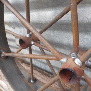 May include: Close-up of a rusty metal wheel with spokes and a central hub. The spokes are welded to the hub with visible weld marks. The wheel is made of metal and has a rustic appearance.