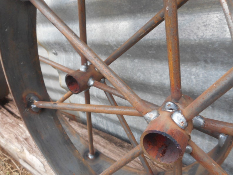 May include: Close-up of a rusty metal wheel with spokes and a central hub. The spokes are welded to the hub with visible weld marks. The wheel is made of metal and has a rustic appearance.