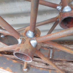 Puede incluir: Primer plano de una rueda de metal oxidada con radios y un cubo. El cubo est&aacute; hecho de un tubo de metal grueso con un agujero en el centro. Los radios est&aacute;n soldados al cubo y a la llanta. La rueda est&aacute; parcialmente cubierta de &oacute;xido.
