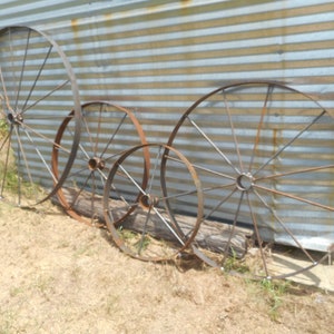 May include: Four rusty metal wagon wheels of varying sizes. The wheels are arranged in a row on a dirt surface in front of a corrugated metal wall.