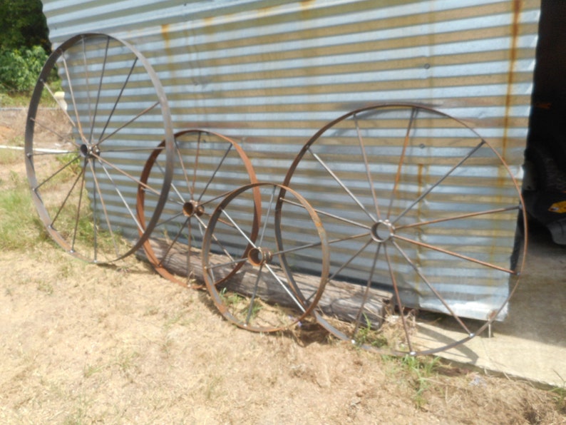 May include: Four rusty metal wagon wheels of varying sizes. The wheels are arranged in a row on a dirt surface in front of a corrugated metal wall.