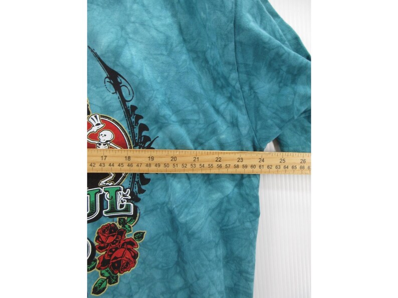 May include: Teal tie-dye t-shirt with a graphic featuring a skeleton, roses, and the word "SOUL". The design includes black and red accents. A wooden ruler is placed across the shirt, measuring approximately 26 inches.