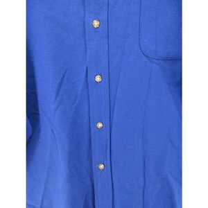 May include: A solid royal blue button-down shirt featuring a vertical placket with four visible light-colored circular buttons and a single chest pocket on the right side. The fabric has a smooth texture and a consistent deep blue color throughout.