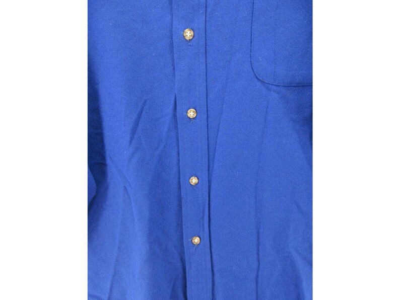 May include: A solid royal blue button-down shirt featuring a vertical placket with four visible light-colored circular buttons and a single chest pocket on the right side. The fabric has a smooth texture and a consistent deep blue color throughout.