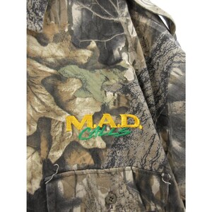 Puede incluir: Camisa con estampado de camuflaje con el texto bordado "MAD CATS" en amarillo y verde. La camisa tiene una parte delantera abotonada y un bolsillo. La tela tiene una mezcla de tonos marrones, verdes y grises.