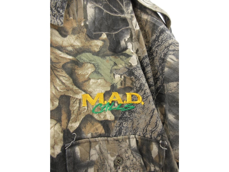 Puede incluir: Camisa con estampado de camuflaje con el texto bordado "MAD CATS" en amarillo y verde. La camisa tiene una parte delantera abotonada y un bolsillo. La tela tiene una mezcla de tonos marrones, verdes y grises.