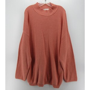 May include: A coral-colored sweater with a mock neck and long sleeves. The sweater is made of a textured knit fabric and hangs on a white hanger against a white background. The sweater has a relaxed fit.