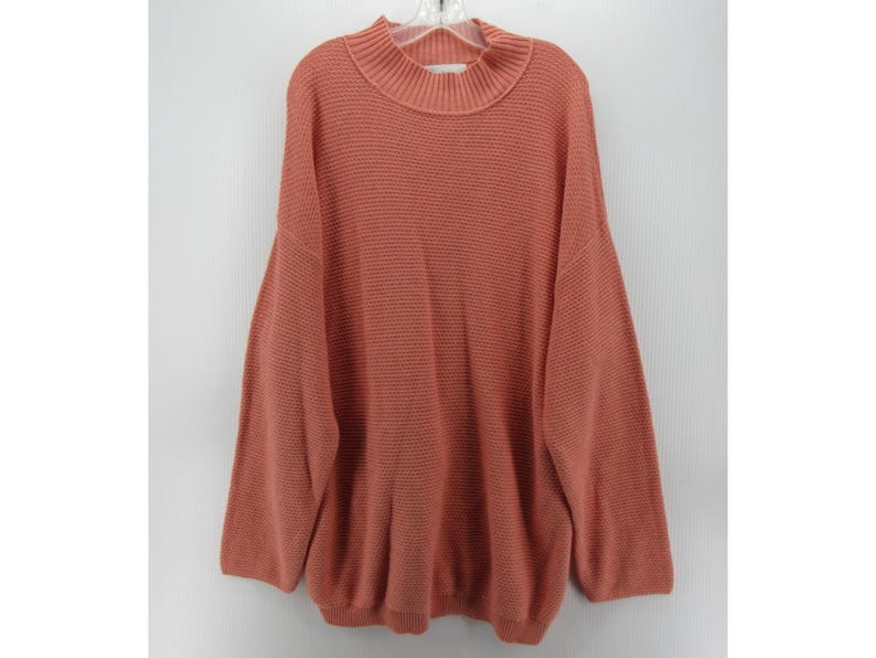 May include: A coral-colored sweater with a mock neck and long sleeves. The sweater is made of a textured knit fabric and hangs on a white hanger against a white background. The sweater has a relaxed fit.