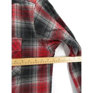 May include: A long-sleeve button-up shirt featuring a red, black, and grey plaid pattern. A wooden ruler is placed across the chest, showing a measurement of approximately 68.5 cm from the side seam to the centre of the shirt front.