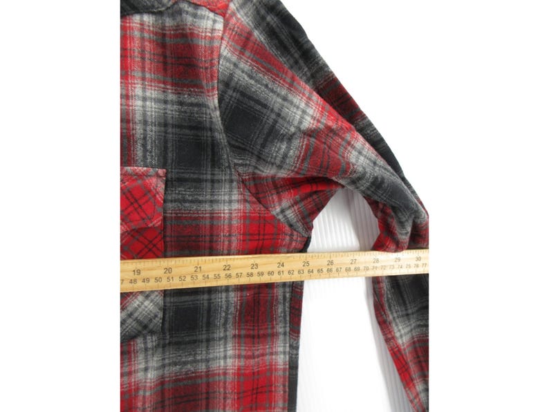 May include: A long-sleeve button-up shirt featuring a red, black, and grey plaid pattern. A wooden ruler is placed across the chest, showing a measurement of approximately 68.5 cm from the side seam to the centre of the shirt front.