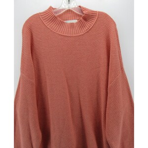 May include: A coral-colored sweater with a mock neck and a textured knit pattern. The sweater has long sleeves and a relaxed fit. The garment is hanging on a white hanger against a white background.