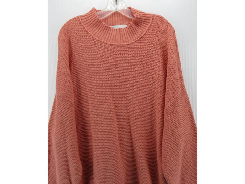 May include: A coral-colored sweater with a mock neck and a textured knit pattern. The sweater has long sleeves and a relaxed fit. The garment is hanging on a white hanger against a white background.