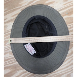 Op de afbeelding: Olijfgroene zonnehoed met een zwarte binnenkant en een brede rand. Een houten liniaal is over de binnenkant van de hoed geplaatst, met een breedte van ongeveer 28 cm. De hoed heeft een label aan de binnenkant.