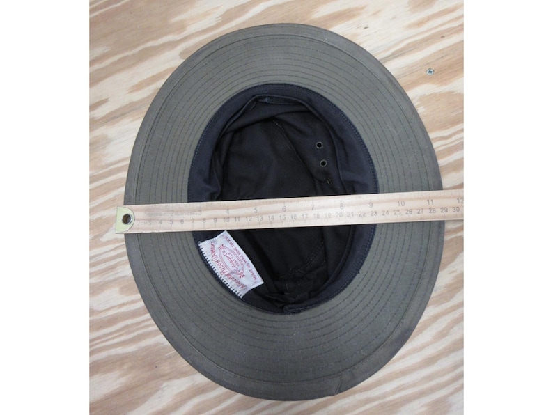 Op de afbeelding: Olijfgroene zonnehoed met een zwarte binnenkant en een brede rand. Een houten liniaal is over de binnenkant van de hoed geplaatst, met een breedte van ongeveer 28 cm. De hoed heeft een label aan de binnenkant.