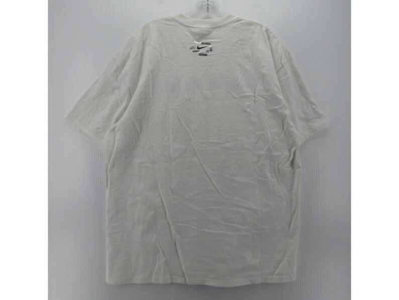 May include: A white short-sleeved t-shirt with a small logo near the neckline. The shirt is hanging on a hanger against a plain white background. The fabric appears to be slightly wrinkled.