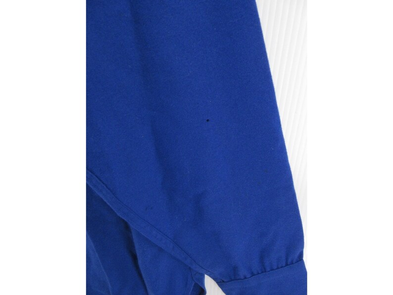 May include: A long-sleeved shirt in a solid, vibrant royal blue color. The garment features a smooth, opaque fabric texture with a visible cuff at the end of the sleeve and a small, dark spot imperfection on the upper portion of the fabric.