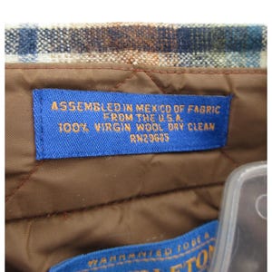 May include: A brown quilted lining inside a garment with a blue rectangular label featuring orange text that reads: ASSEMBLED IN MEXICO OF FABRIC FROM THE U.S.A. 100% VIRGIN WOOL DRY CLEAN RN29685. Above the label is a blue, brown, and white plaid fabric.