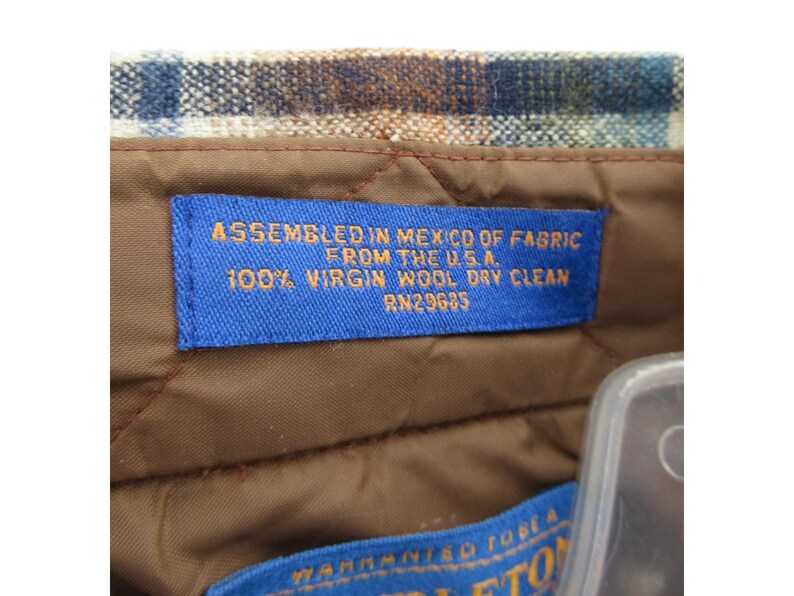 May include: A brown quilted lining inside a garment with a blue rectangular label featuring orange text that reads: ASSEMBLED IN MEXICO OF FABRIC FROM THE U.S.A. 100% VIRGIN WOOL DRY CLEAN RN29685. Above the label is a blue, brown, and white plaid fabric.